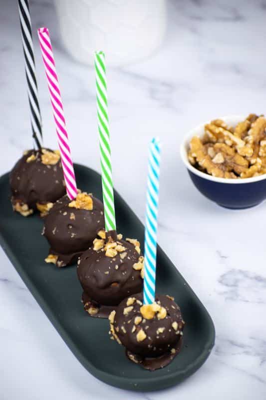 Banana Cake Pops_6 Banana cake pops upside down on a black tray with colorful, striped treat sticks. Walnuts in a small dish on the side.