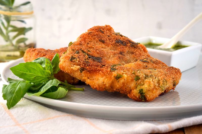 Electric skillet fried fish on a plate with basil pesto in the background and basil leaves on the dish.