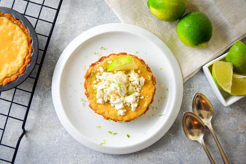 Mini Electric Skillet Lime Pie on a white plate, limes and spoons on the side, on grey background.