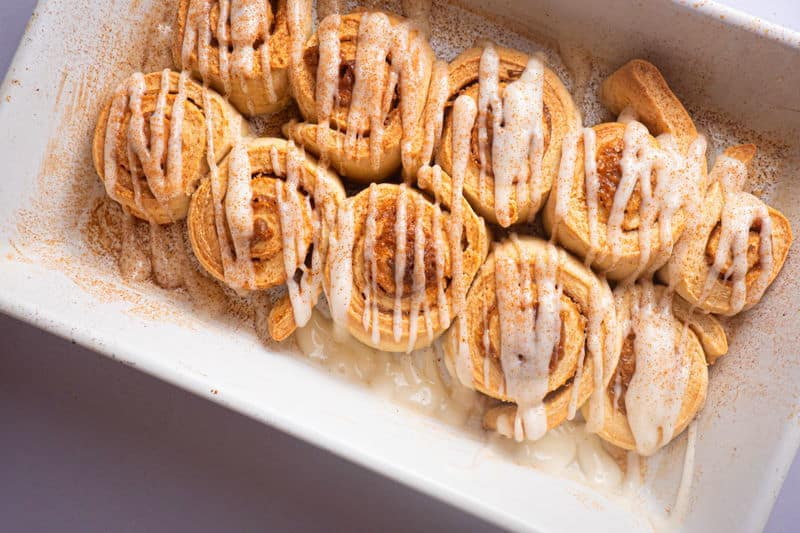 How to Make Cinnamon Rolls_24 Cinnamon Buns with cream cheese frosting drizzled on top in white serving dish.