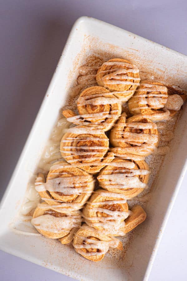 How to Make Cinnamon Rolls_25 Cinnamon Buns with cream cheese frosting drizzled on top in white serving dish.