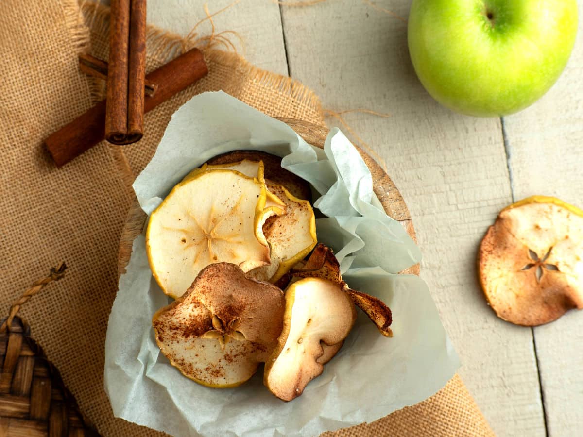 Cinnamon apple chips in a parchment-lined wooden bowl with cinnamon sticks on the side.