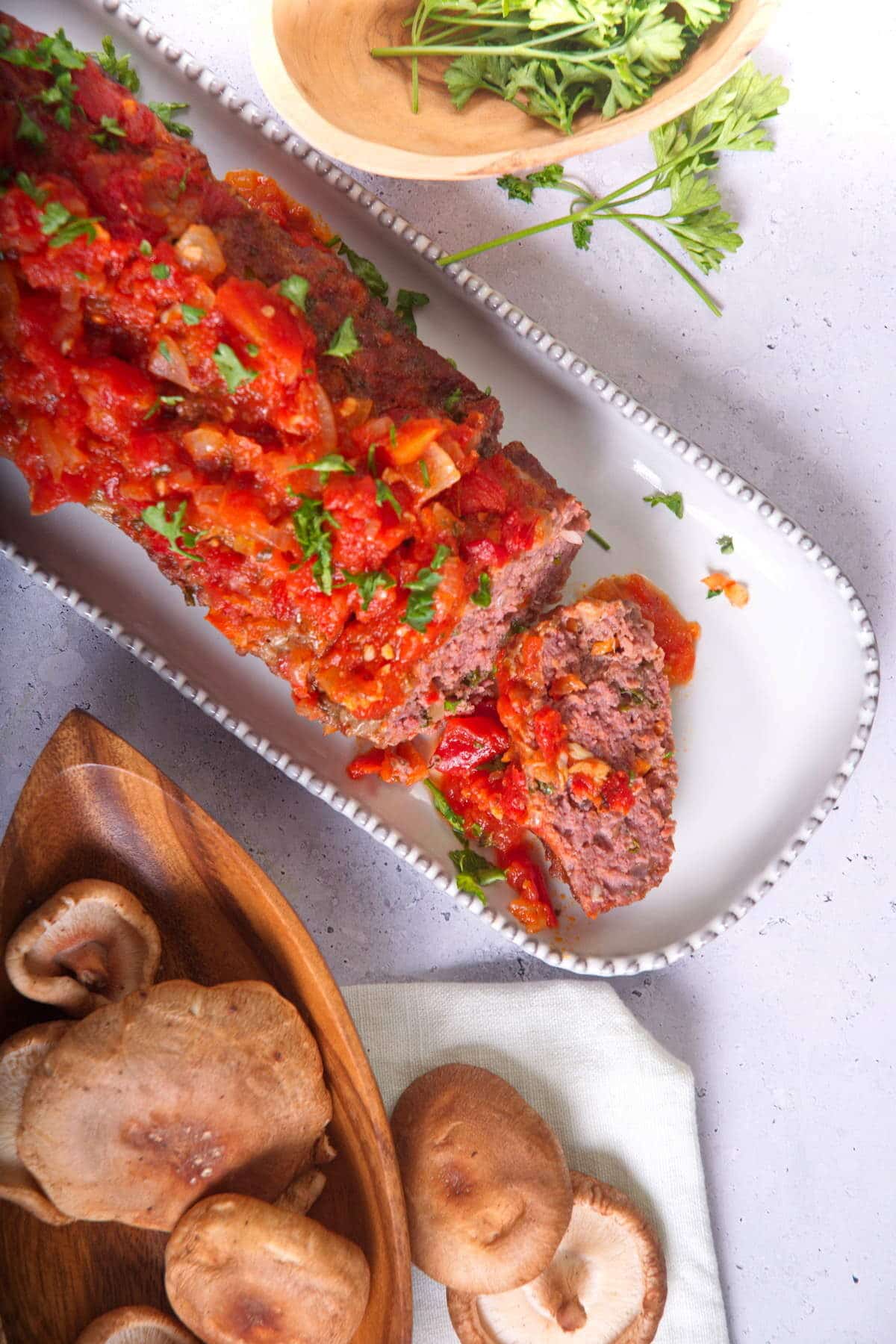 Bison meatloaf with marinara sauce sliced on white serving dish, mushrooms and parsley on the side.