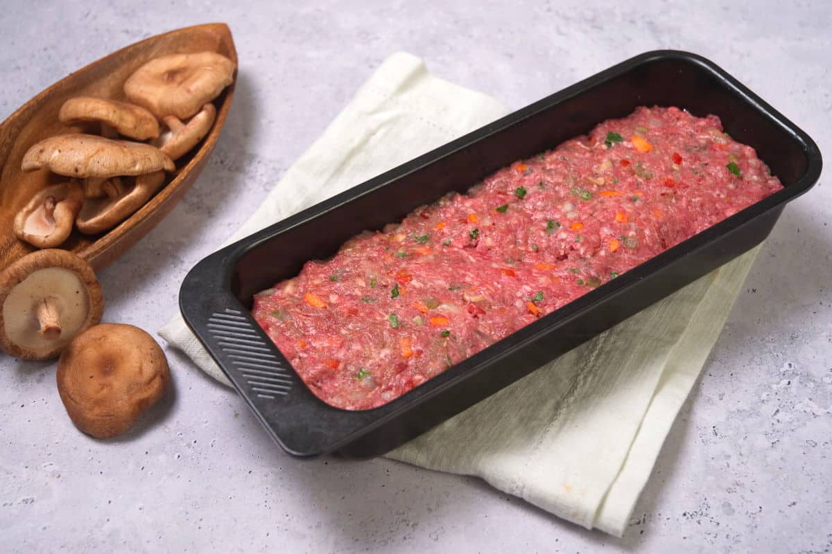 Raw bison meatloaf in loaf pan with mushrooms on the side.