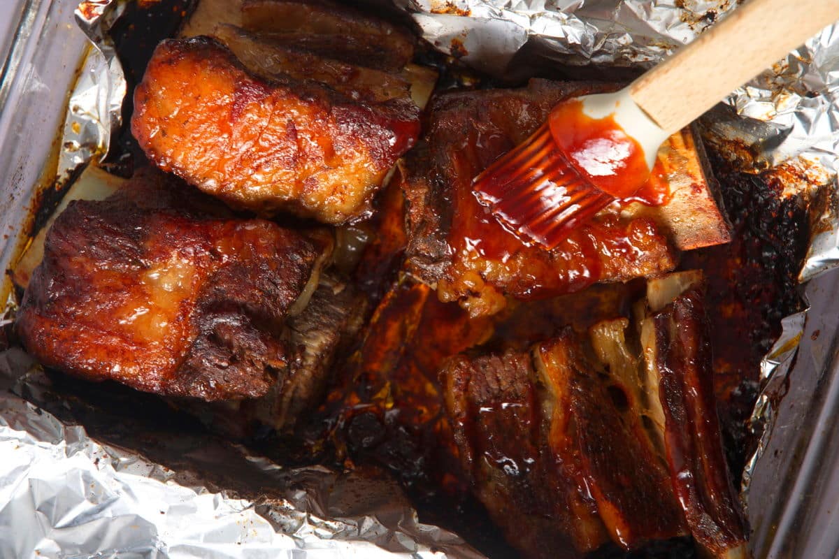 Bison short ribs getting covered with BBQ sauce in foil-lined dish.