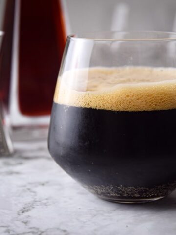 Black fog cocktail in a glass, raspberry liqueur in the background, on marble.