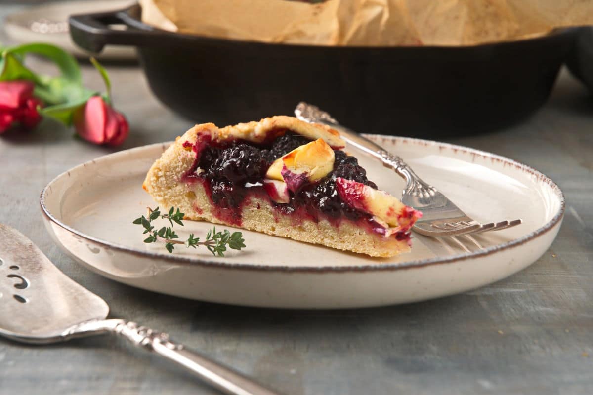 Slice of blackberry galette on plate with fork and thyme sprig.