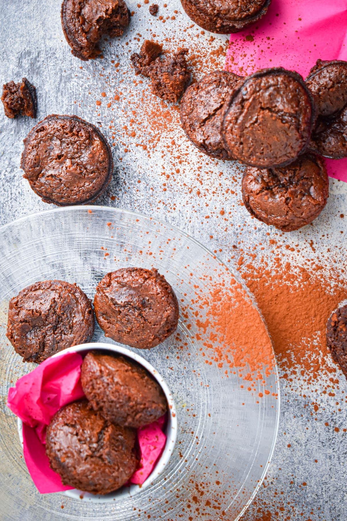Brownie bites scattered on and around clear plate with cocoa powder and pink napkins.