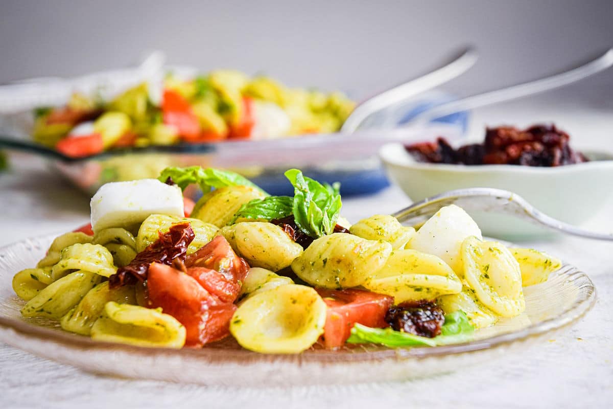Caprese pasta salad on clear glass plate and small bowl of sun-dried tomatoes in the background.