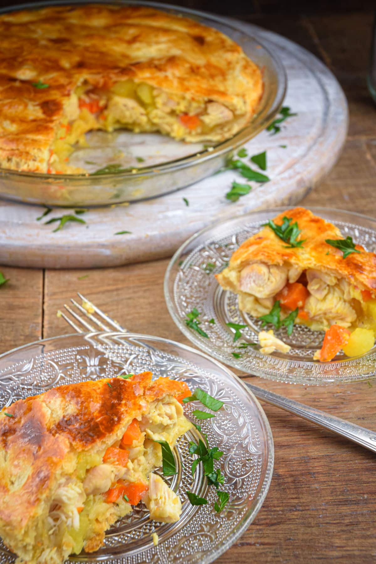 Chicken pasty pie in pie dish and on clear glass plates on brown wooden background.