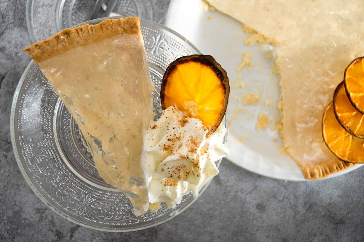 Clear pumpkin pie slice on a clear glass plate with dehydrated orange slices and whipped cream.