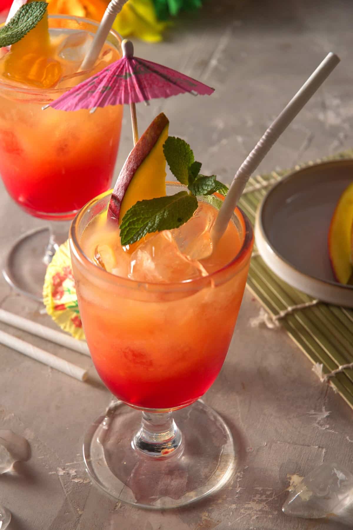 Mai tais in cocktail glasses with umbrella, mango slice, mint sprig and straw on concrete-style background.