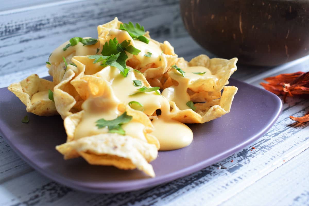 Tortilla chips covered in white nacho cheese sauce on a purple plate, white wooden background.