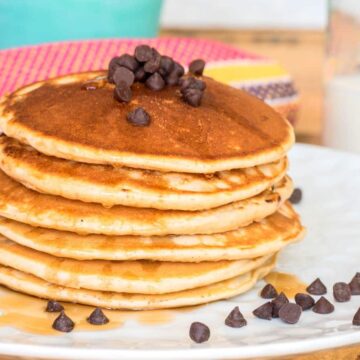 Pancakes piled on a plate with chocolate chips and maple syrup.