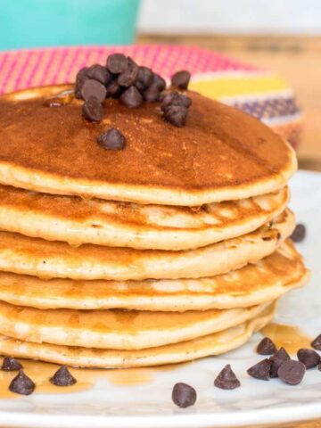 Pancakes piled on a plate with chocolate chips and maple syrup.