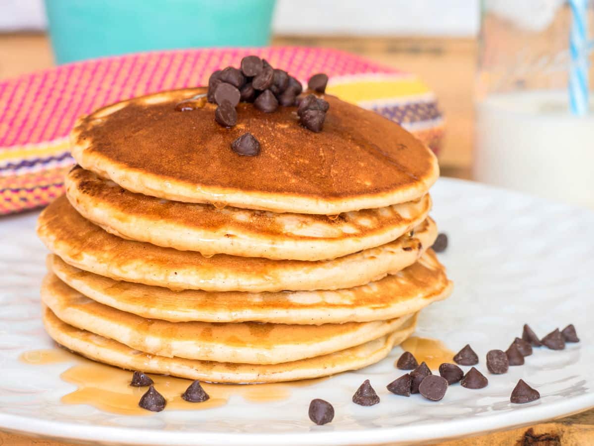 Pancakes piled on a plate with chocolate chips and maple syrup.