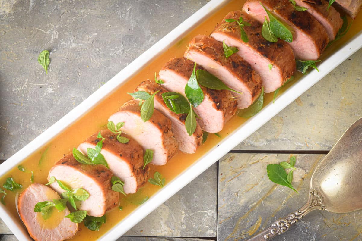 Sliced pork tenderloin in white serving dish with orange white wine sauce.