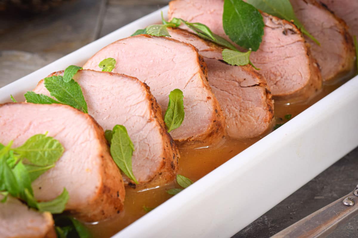 Sliced pork tenderloin in white serving dish with orange white wine sauce and fresh herbs.