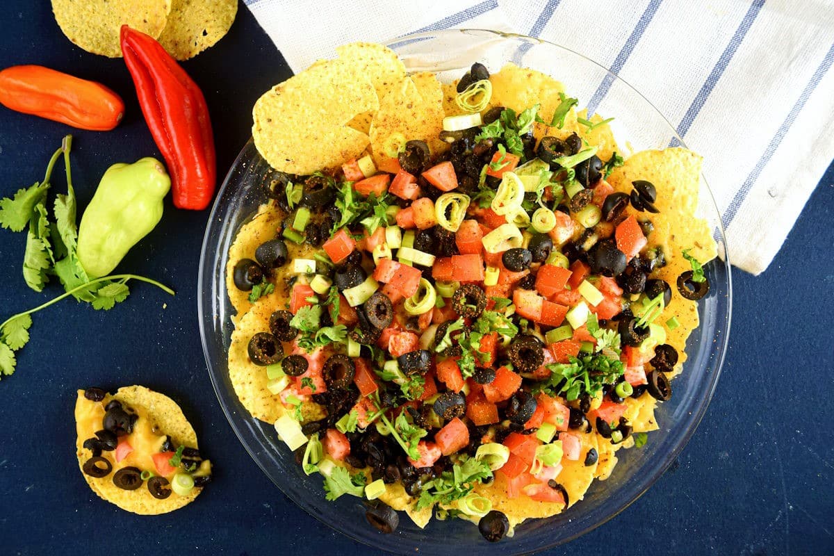 Veggie nachos on a clear glass plate on blue background with peppers on the side.