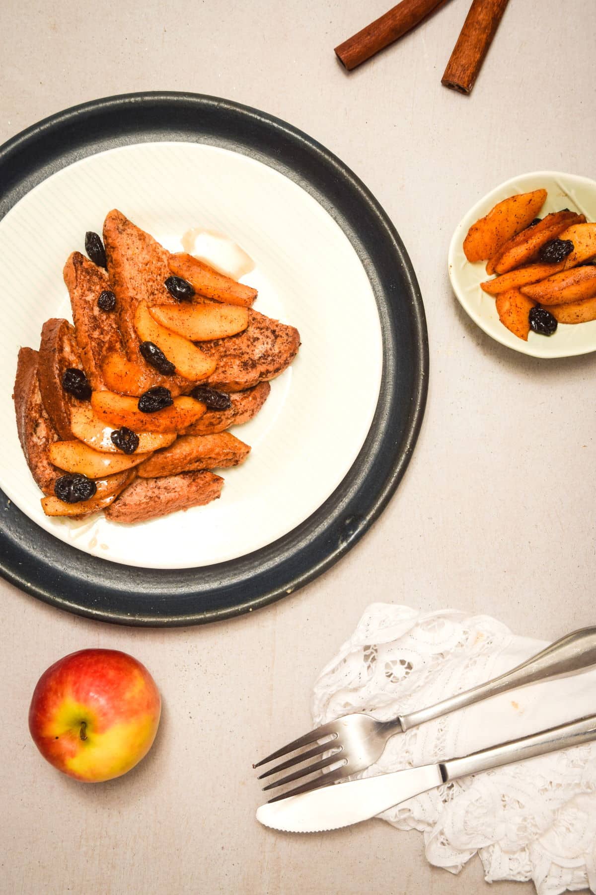 French toast with sliced apples and raisins on white and black plate. Fork and knife on the side.