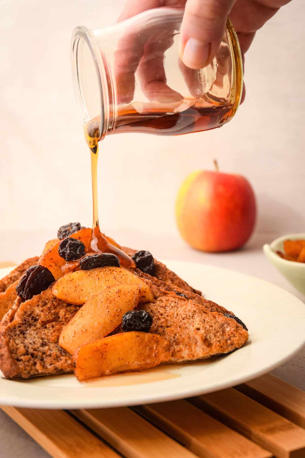 French toast with sliced apples and raisins on white plate and maple syrup pouring over top.