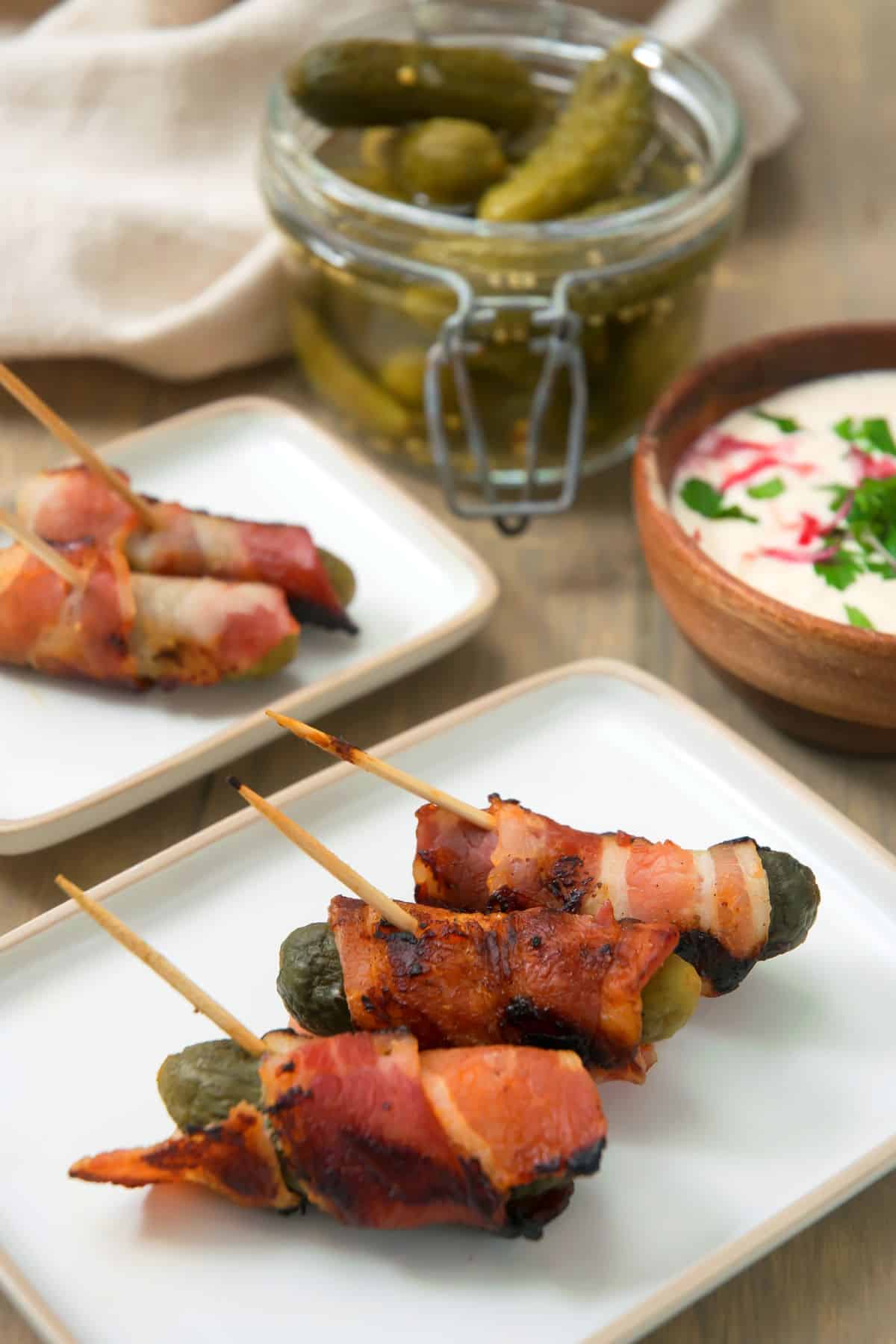 Grilled, bacon wrapped pickles on toothpicks, on a white plate, with dipping sauce in the background.