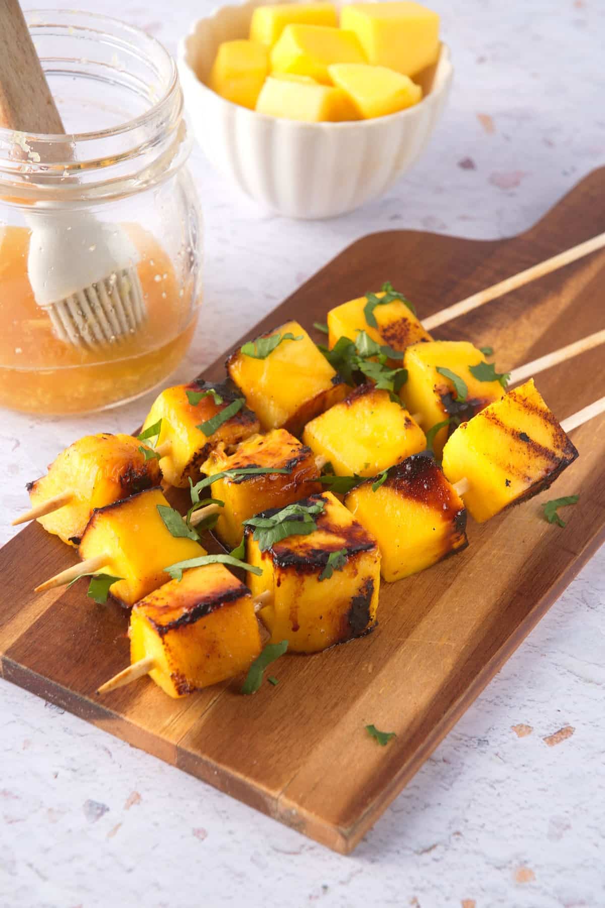 Grilled mango skewers on wooden serving board with fresh cilantro and marinade on the side.