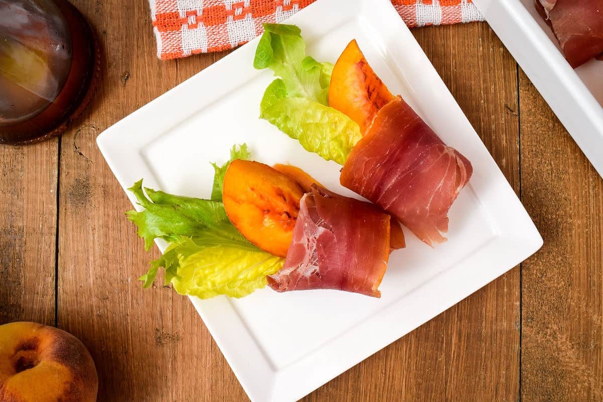 Grilled Peaches Wrapped in Proscuitto 5 Grilled Peaches Wrapped in Proscuitto on a porcelain appetizer plate, balsamic dressing in the background.