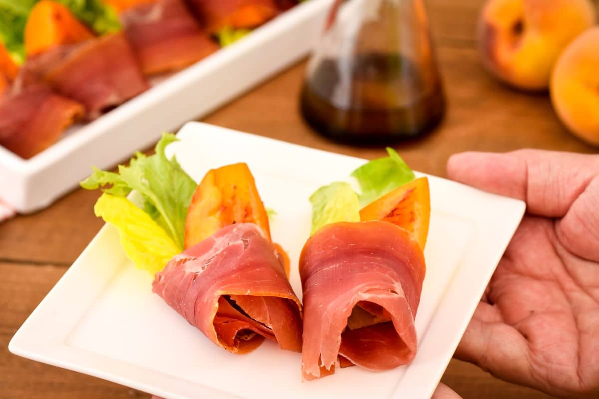 Grilled Peaches Wrapped in Proscuitto on a porcelain appetizer plate with hand holding it.