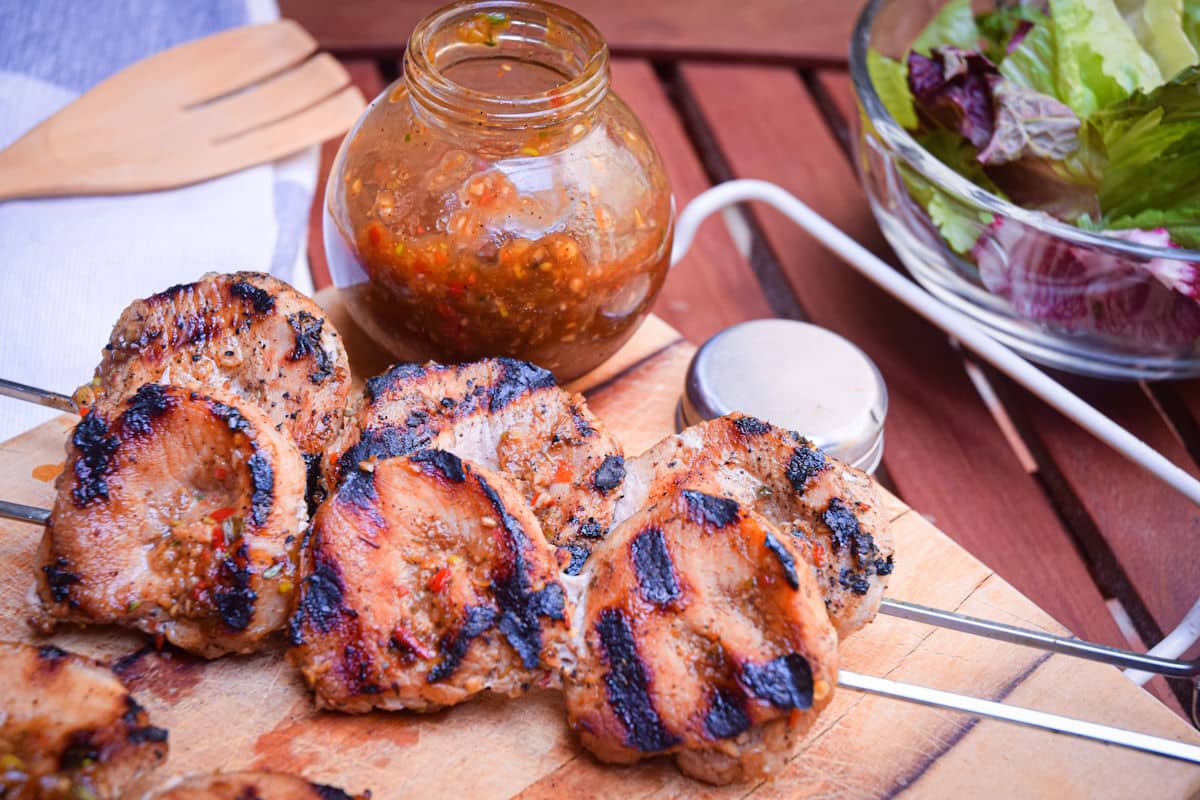 Grilled pork skewers on wooden cutting board with salad and jerk sauce on the side.