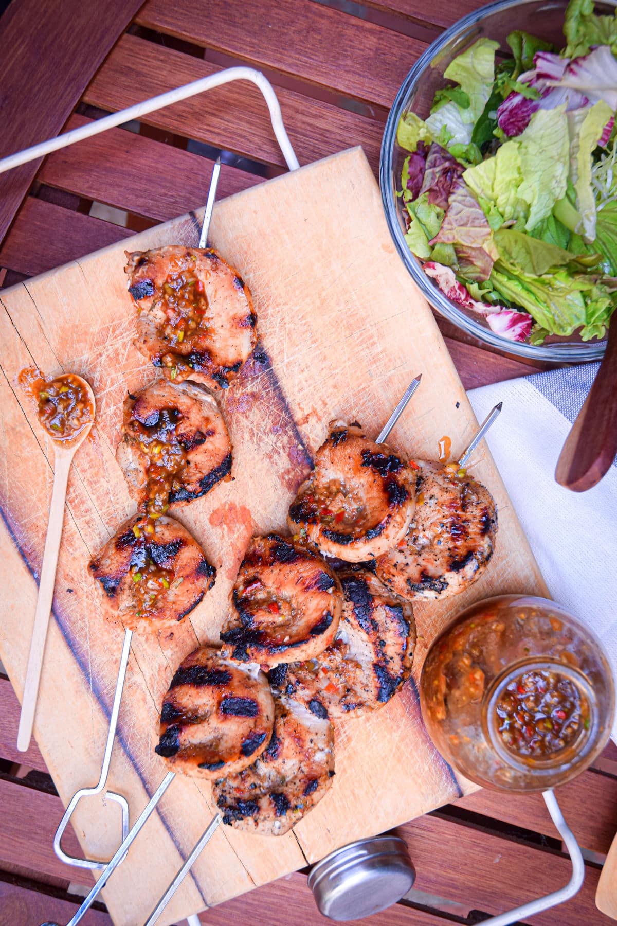 Grilled pork skewers on wooden cutting board with salad and jerk sauce on the side.