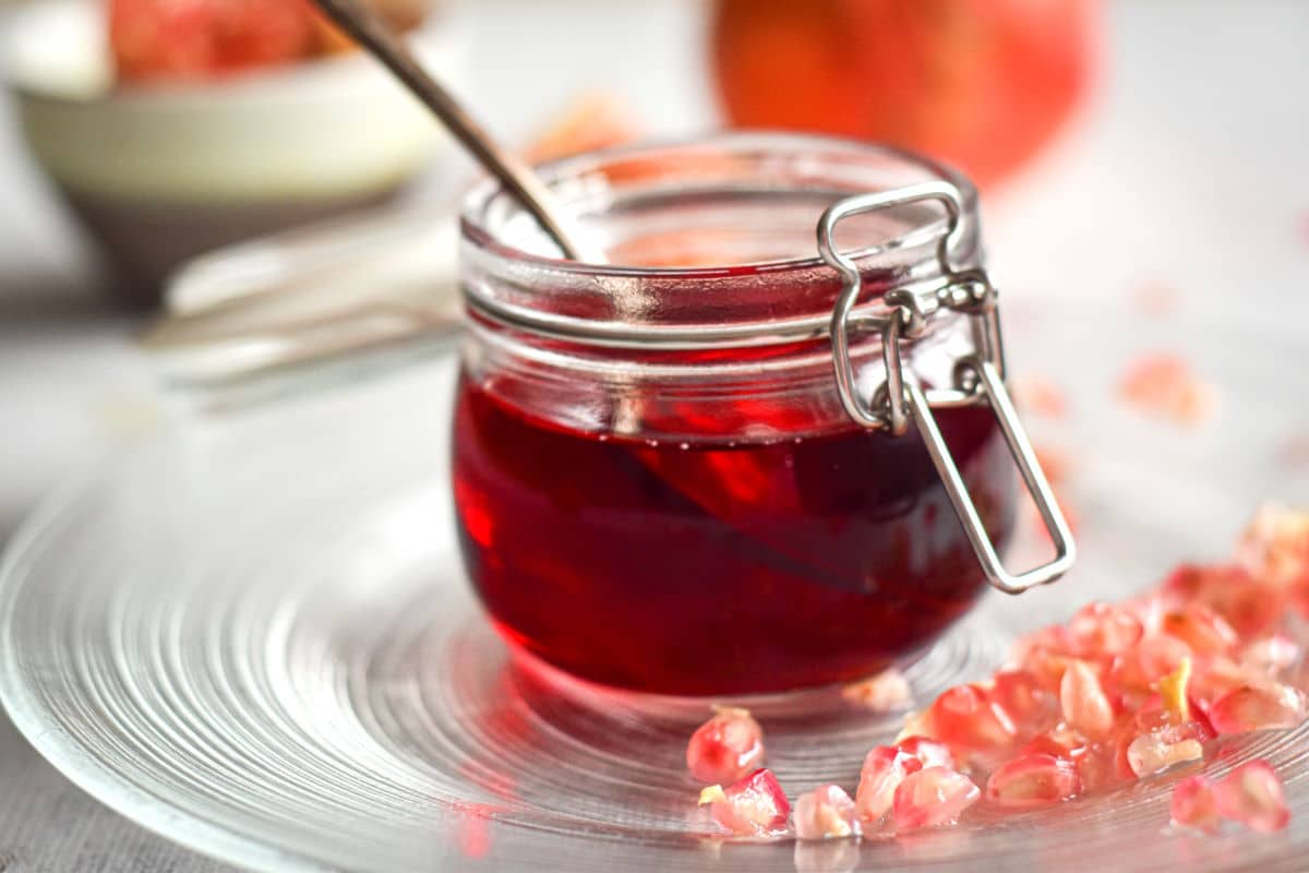 Homemade grenadine syrup in a small glass jar surrounded by pomegranate seeds.