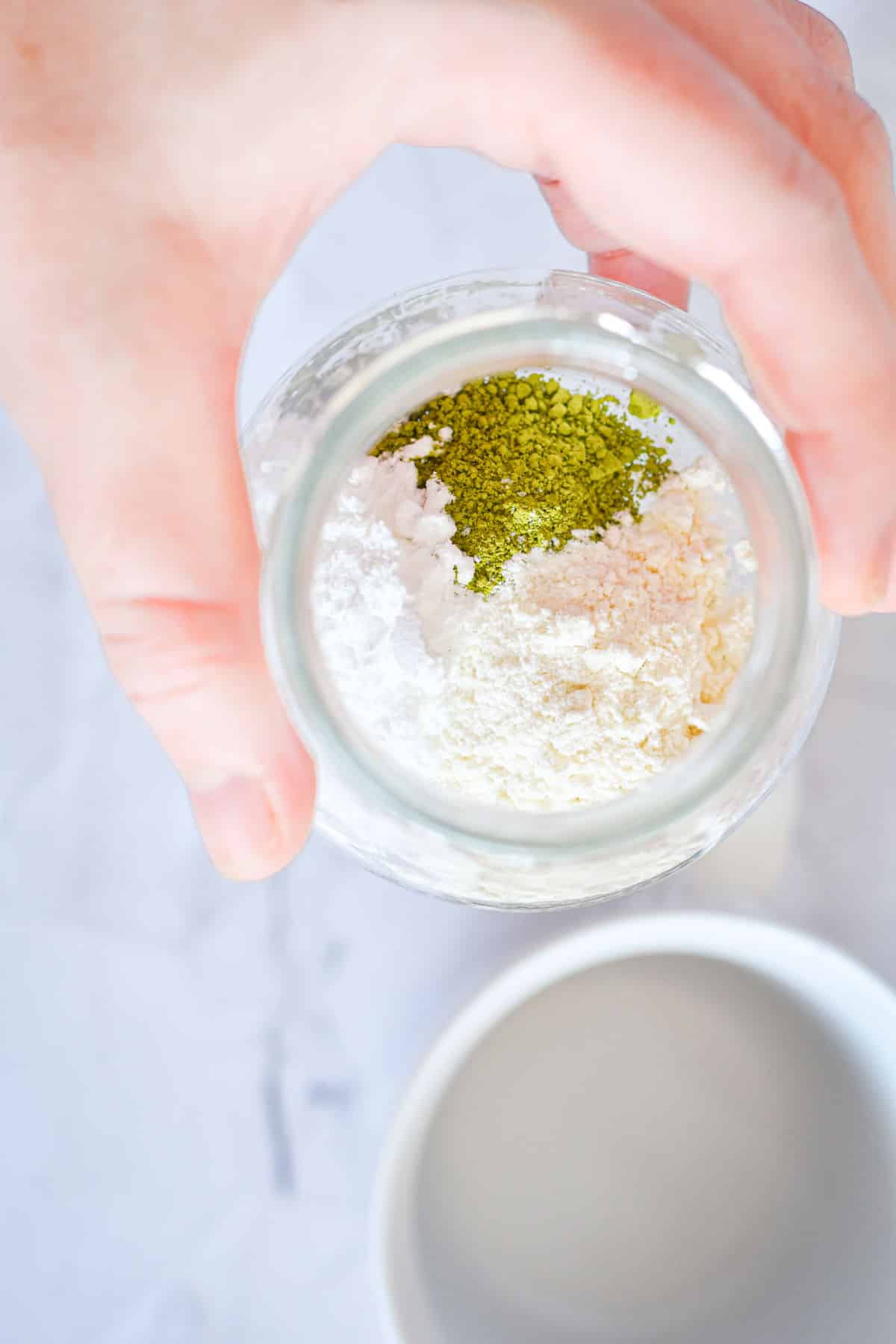A hand holding vegan matcha latte powder in small jar unmixed.