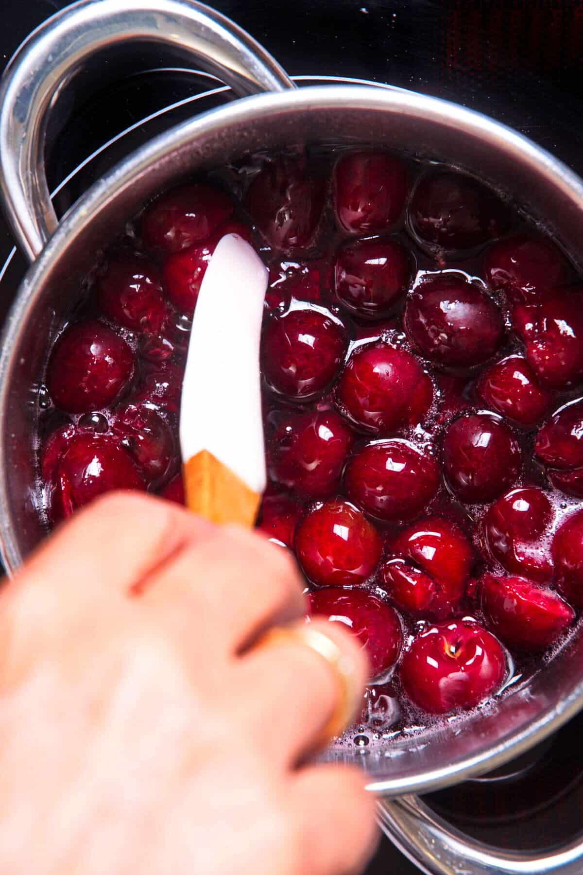 Cherries in a sugar syrup getting stirred with spatula in saucepan.