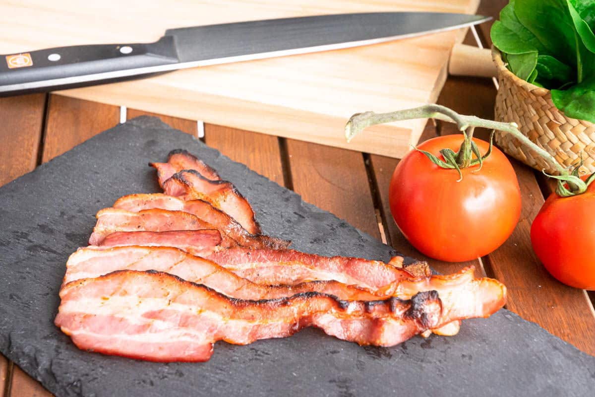 Crispy bacon on a black board, with fresh tomatoes and knife on the side.