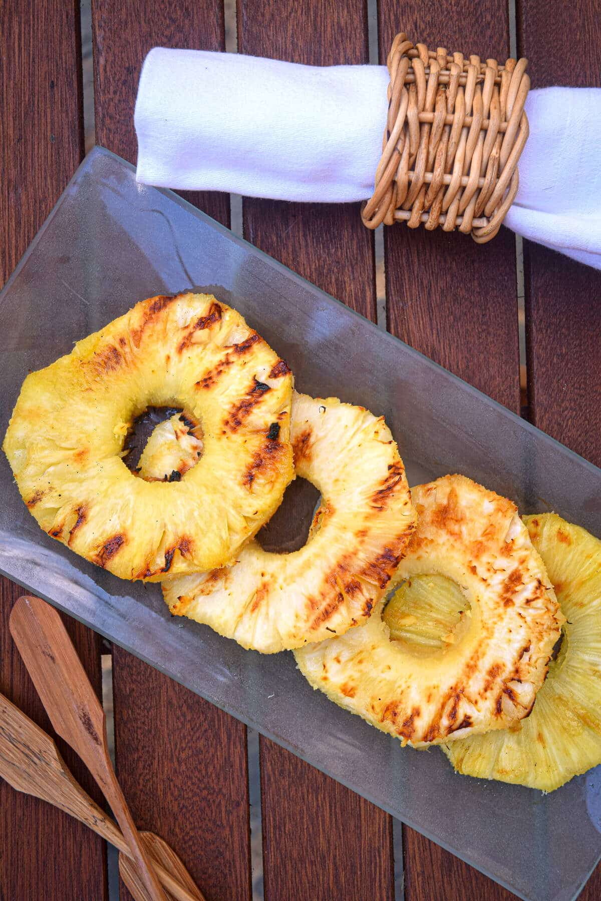 How to Grill Pineapple Slices_17 Grilled pineapple slices on glass serving dish and a cloth napkin on wooden table.