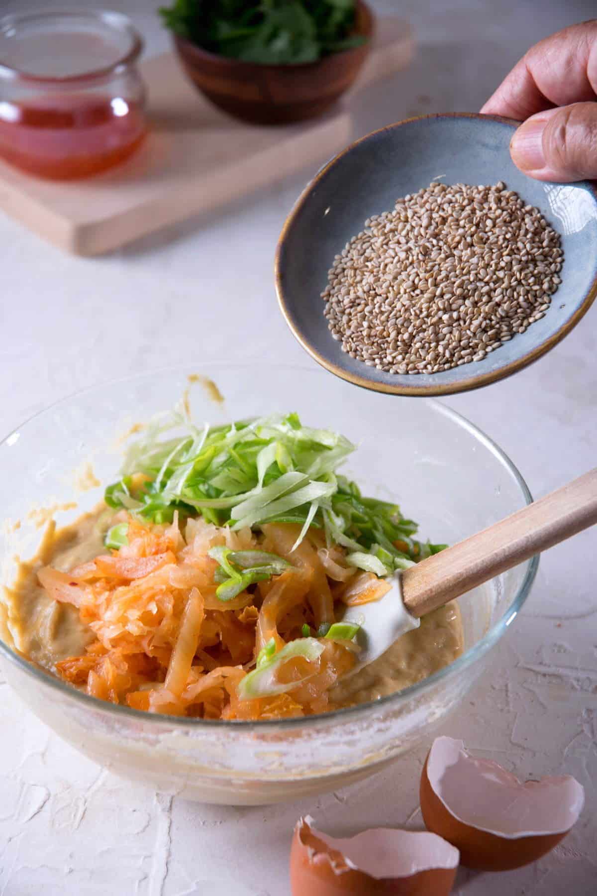 Kimchi pancake ingredients in a glass bowl and sesame seeds on a small plate.