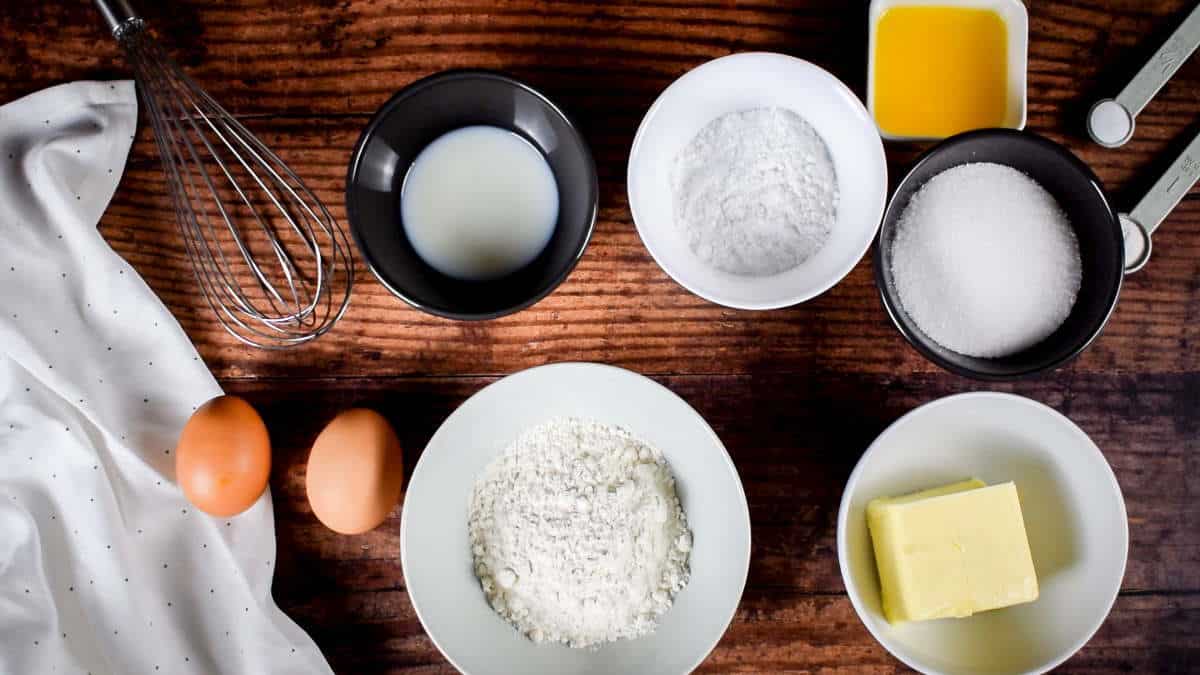 How to Make Cake Pops with a Cake Pop Maker_Prep Cake pop ingredients in bowls on wooden background, as well as a whisk.