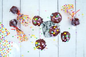 Chocolate cake pops with sprinkles, wrapped in cellophane, on white wooden background.