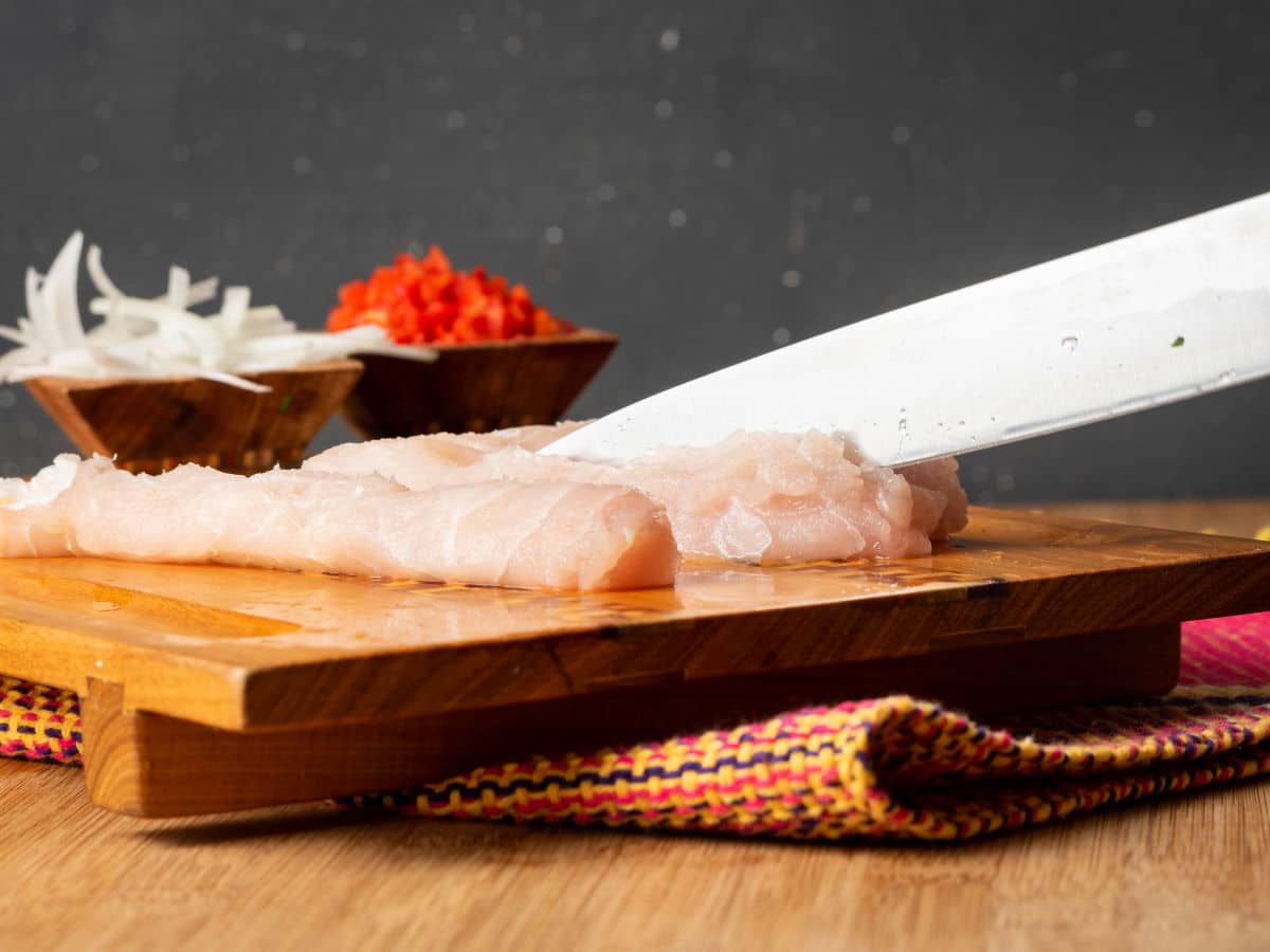 How to Make Ceviche - Step 2 A knife slicing into raw white fish on cutting board.