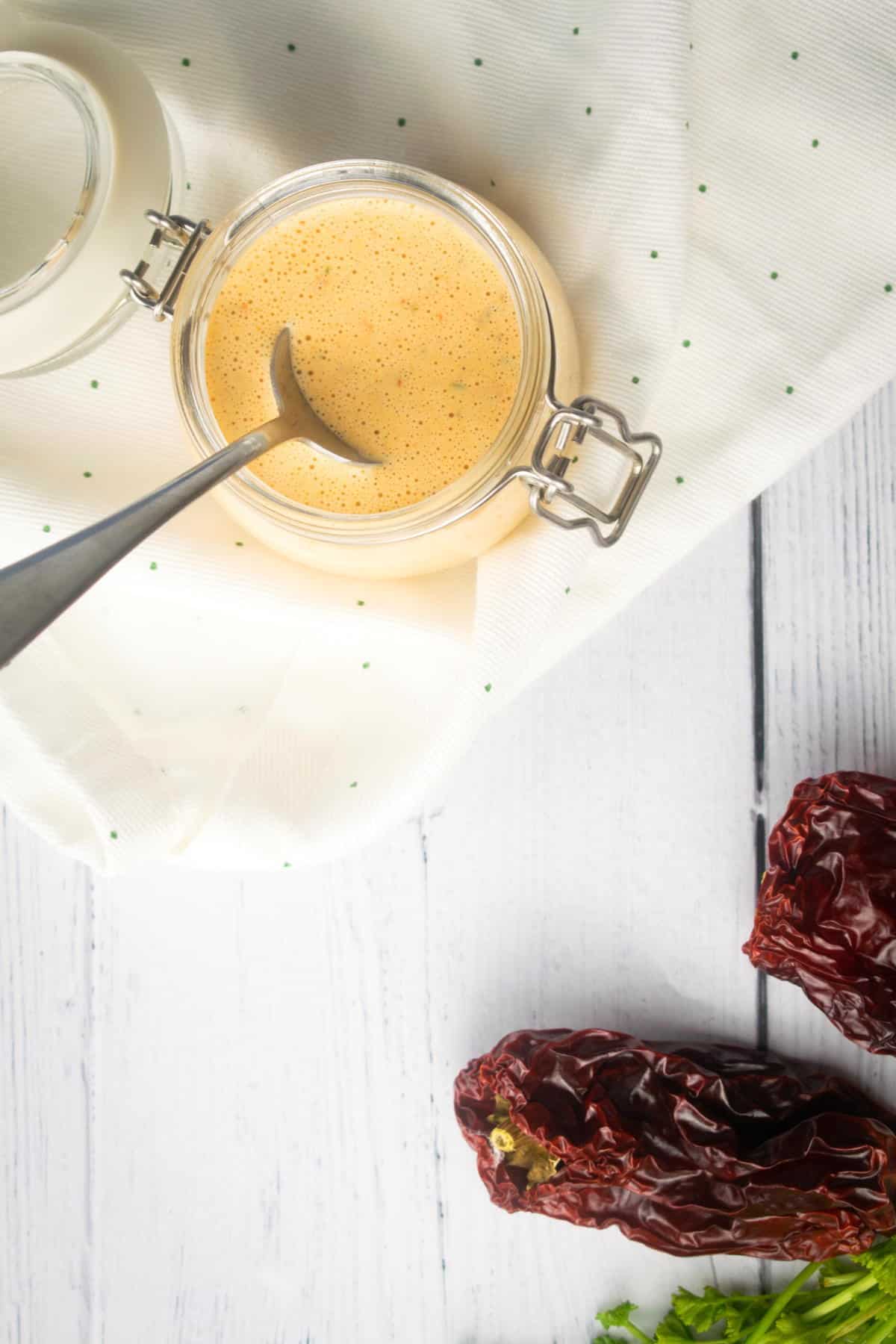 How to make chipotle sauce Step 3-5 Chipotle sauce in a jar with a spoon on white wooden background. Chipotle peppers on the side.