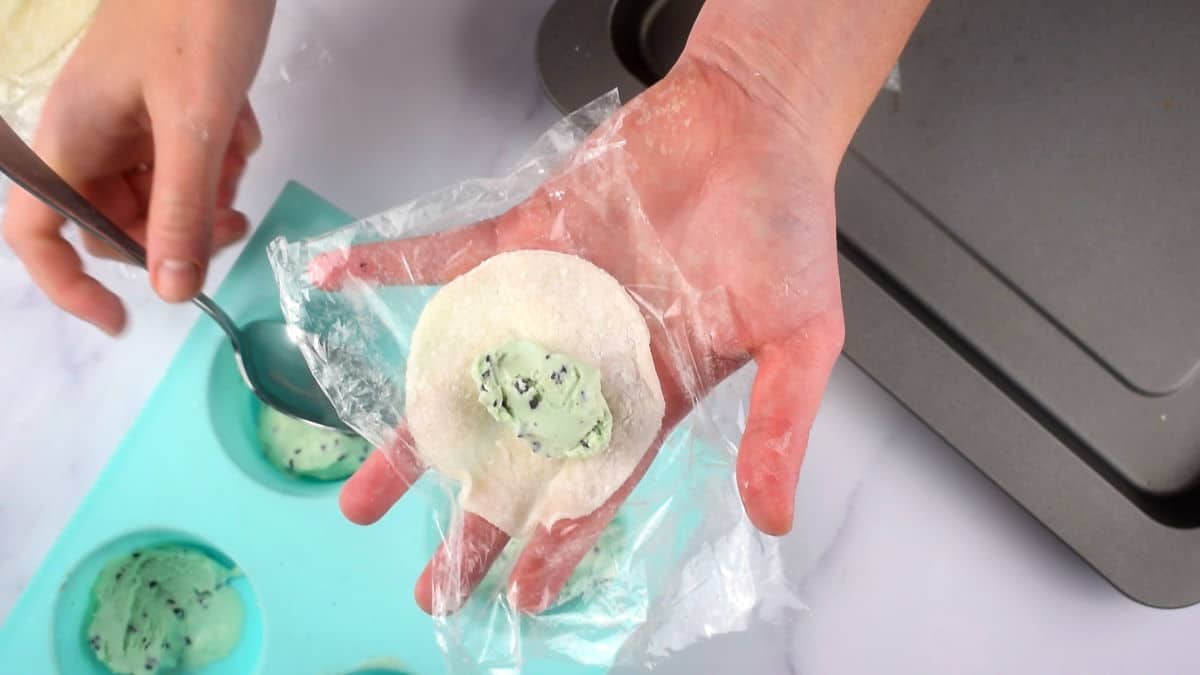 How to Make Mochi Ice Cream Step 7-2 Mochi dough circle with mint chocolate chip ice cream on plastic wrap in a hand.