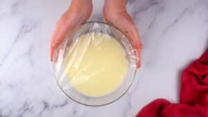 Flour mixture in small glass bowl with plastic wrap on top, on marble background.