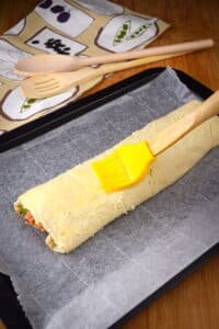 Unbaked sausage roll and pastry brush on baking sheet.