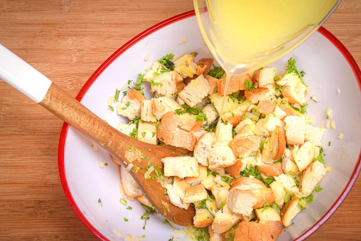 Raw stuffing mix in a bowl with a wooden spoon and glass of chicken broth.