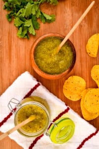 A wooden bowl of tomatillo salsa, a jar of tomatillo salsa, cilantro and tortilla chips in the background.