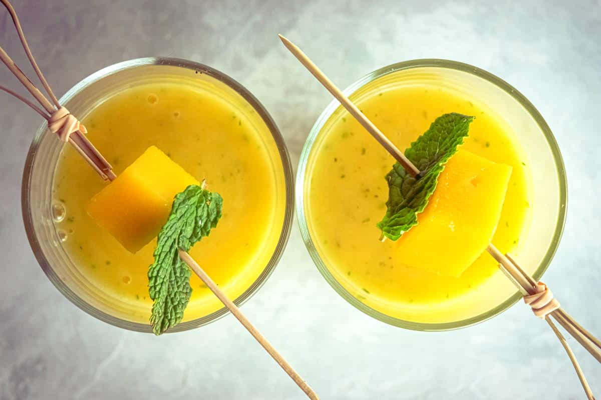 Mango pineapple smoothies in 2 glasses with mango and mint skewers.