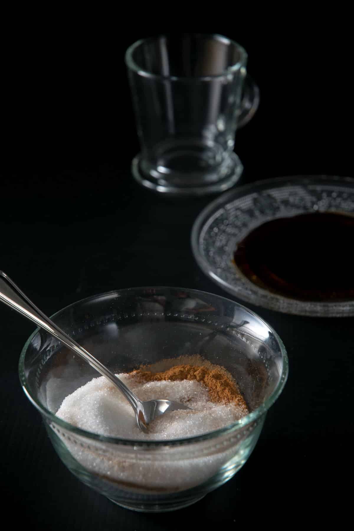 Cinnamon and sugar in a small glass bowl with a spoon on black background.