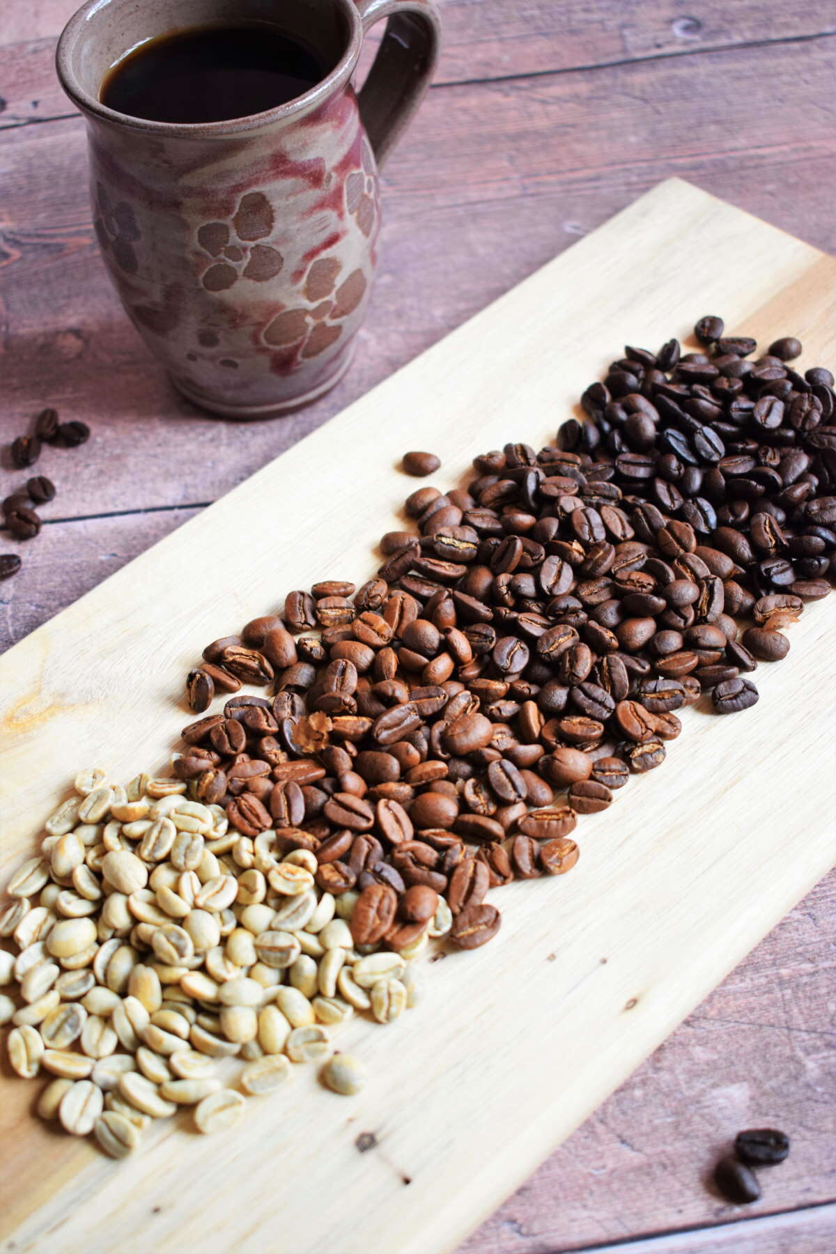 Green and brown coffee beans on wooden serving board, coffee mug in the background.
