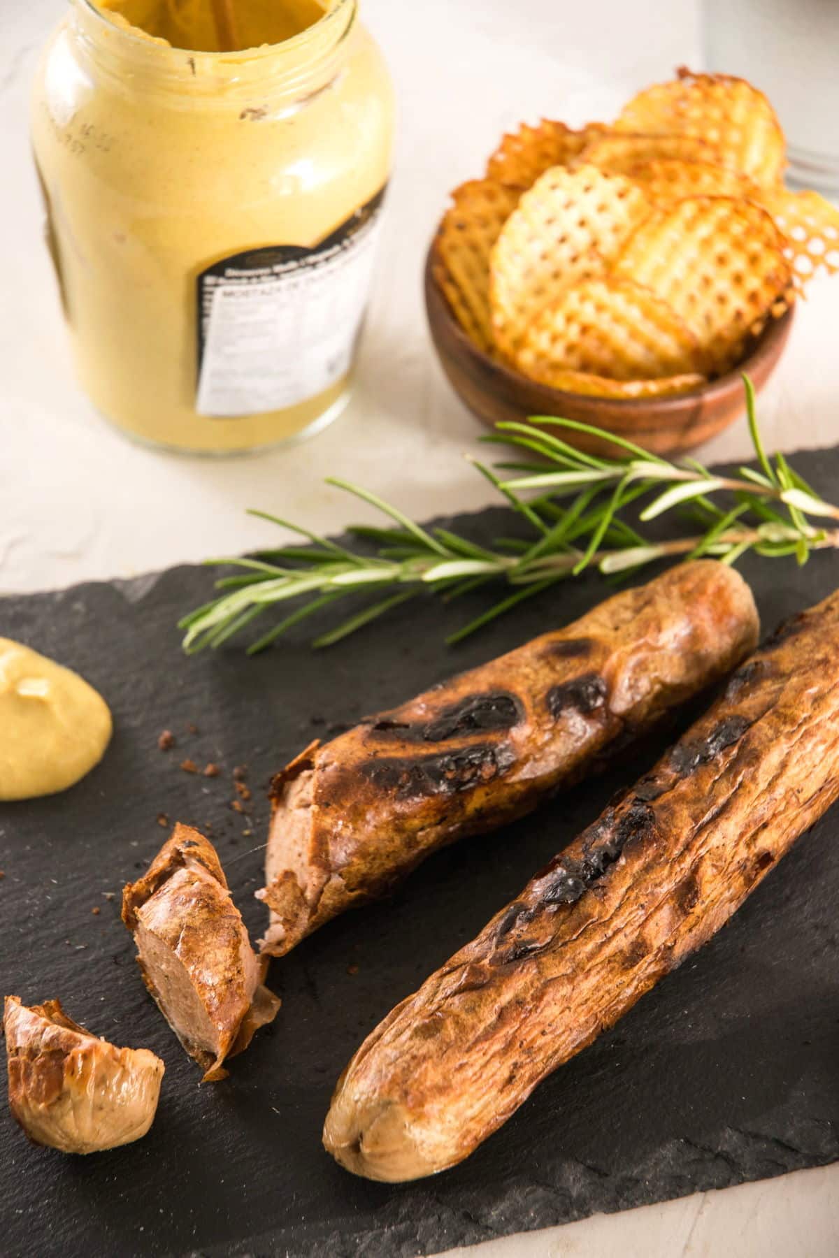 Grilled bratwurst on charcoal cutting board. Rosemary, chips and condiments in the background.